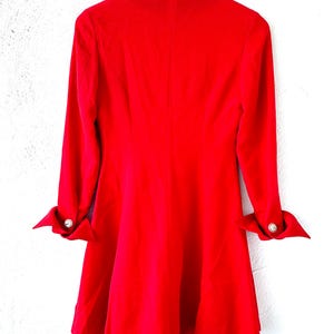 Puede incluir: Un vestido rojo vibrante con mangas largas y falda acampanada, colgado de una percha met&aacute;lica. Las mangas est&aacute;n abotonadas, adornadas con un bot&oacute;n plateado. El vestido es de tela lisa.