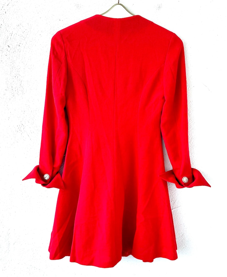 Puede incluir: Un vestido rojo vibrante con mangas largas y falda acampanada, colgado de una percha met&aacute;lica. Las mangas est&aacute;n abotonadas, adornadas con un bot&oacute;n plateado. El vestido es de tela lisa.