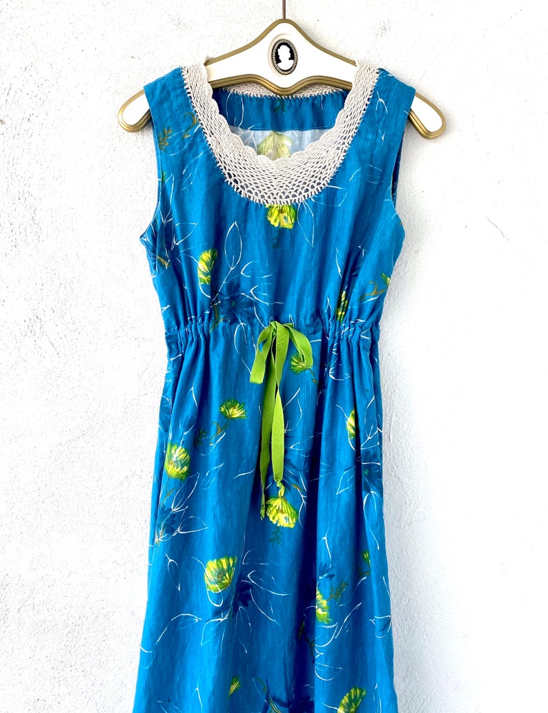 May include: A blue sleeveless dress with a white crochet neckline and a green ribbon tie at the waist. The dress has a floral print with yellow and green flowers.