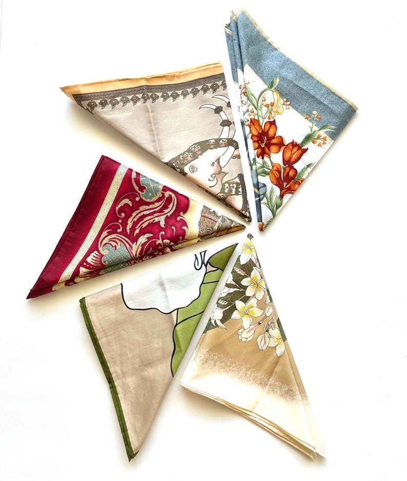 May include: An overhead shot of five folded silk scarves. The scarves feature various patterns, including floral, paisley, and elephant motifs. Colors include red, beige, white, green, and blue. The scarves are arranged in a starburst pattern against a white background.