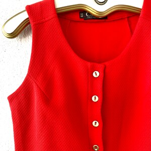 May include: A sleeveless, bright red dress with a textured fabric and a row of gold-colored buttons down the front. The dress is hanging on a gold-colored hanger. The label reads "California".