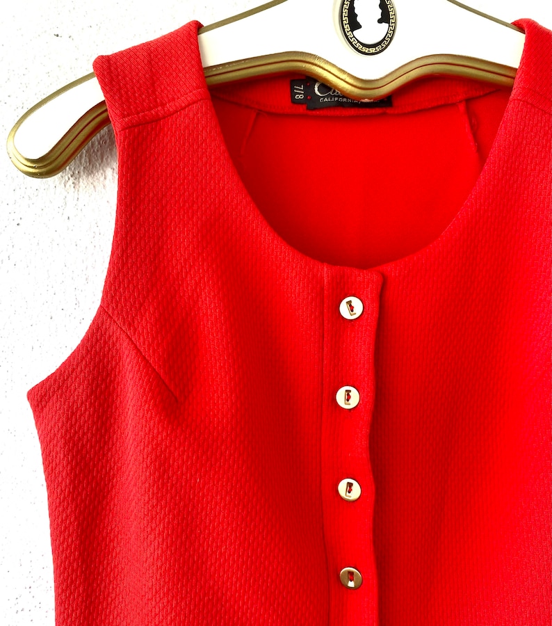 May include: A sleeveless, bright red dress with a textured fabric and a row of gold-colored buttons down the front. The dress is hanging on a gold-colored hanger. The label reads "California".