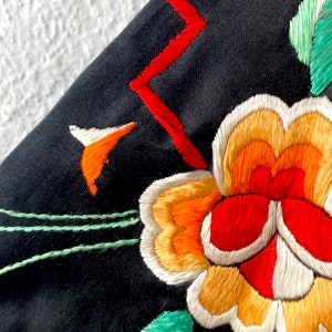 Puede incluir: Primer plano de una tela negra con flores bordadas de colores. Las flores son rojas, naranjas, amarillas y blancas. La tela tiene un dise&ntilde;o bordado verde y rojo.