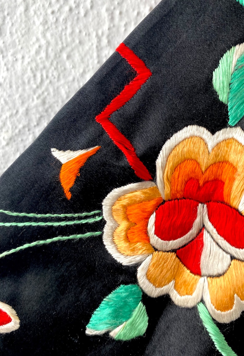 Puede incluir: Primer plano de una tela negra con flores bordadas de colores. Las flores son rojas, naranjas, amarillas y blancas. La tela tiene un dise&ntilde;o bordado verde y rojo.