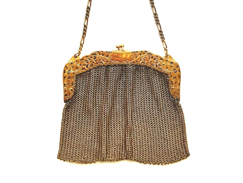 Pu&ograve; includere: Una borsa a rete di catena dorata con una chiusura floreale decorativa. La borsa ha una lunga tracolla a catena.