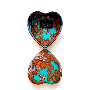 May include: A heart-shaped box with a turquoise and orange floral pattern. The box is open and the inside is lined with the same fabric. The box has a label that says "Diane Von Furstenberg Original".