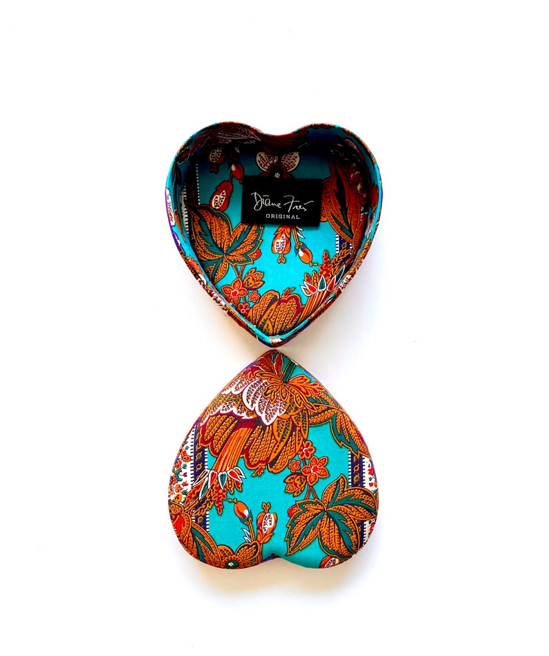 May include: A heart-shaped box with a turquoise and orange floral pattern. The box is open and the inside is lined with the same fabric. The box has a label that says "Diane Von Furstenberg Original".