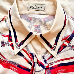 May include: A cream-colored button-down shirt with a collar. The shirt has a red, blue, and white print of equestrian-themed designs. The label on the collar reads "Elan,  Fabrique en Chine".