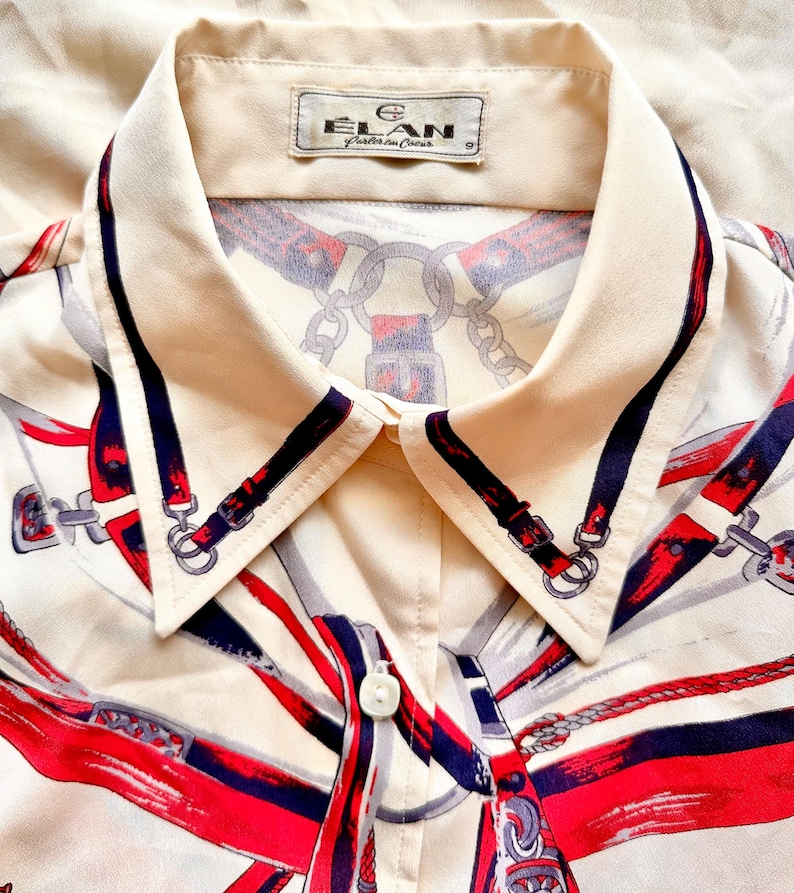 May include: A cream-colored button-down shirt with a collar. The shirt has a red, blue, and white print of equestrian-themed designs. The label on the collar reads "Elan,  Fabrique en Chine".