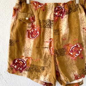 May include: Tan and brown shorts with a floral and geometric pattern. The shorts have a small pocket with a white button. The design includes red and white floral elements and small fish.