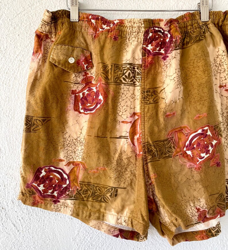 May include: Tan and brown shorts with a floral and geometric pattern. The shorts have a small pocket with a white button. The design includes red and white floral elements and small fish.
