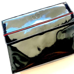 Op de afbeelding: Zwarte lakleren clutch met een rode bies en een zilveren Valentino-logo.