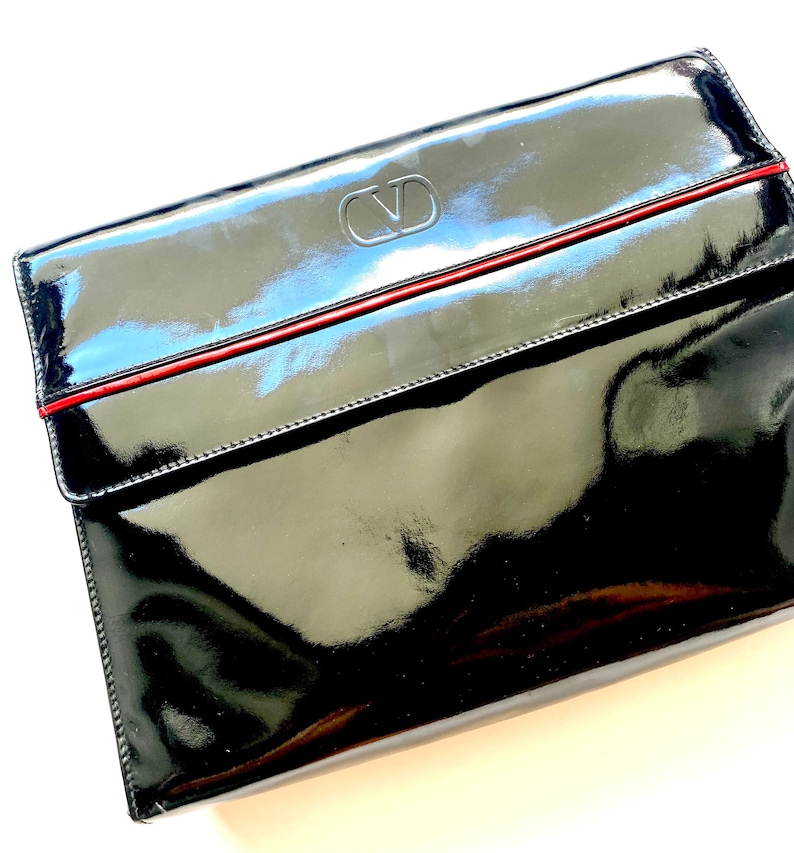 Op de afbeelding: Zwarte lakleren clutch met een rode bies en een zilveren Valentino-logo.