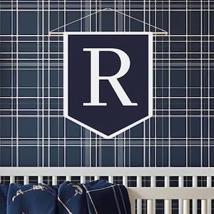Puede incluir: Una cuna blanca con una manta y un juego de sábanas a cuadros azules y blancos. Una pancarta azul marino con la letra "R" cuelga sobre la cuna contra un fondo de papel tapiz a cuadros azul marino.