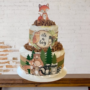 May include: A two-tiered diaper cake decorated with woodland animal figures, including a fox, owl, bear, deer, raccoon, and skunk. The cake is wrapped in burlap ribbon and decorated with green and brown paper shreds. The top tier features a sign that reads "it's a boy".