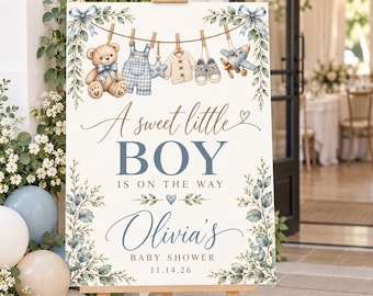 May include: A rectangular sign on a wooden easel featuring a watercolour illustration of a teddy bear, blue overalls, a star, a sweater, shoes, and an aeroplane hanging on a clothesline. Text reads: A sweet little BOY IS ON THE WAY Olivia's BABY SHOWER 14.11.26.