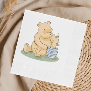 Puede incluir: Servilleta de papel cuadrada blanca con una ilustración de dibujos animados de un oso amarillo sentado en hierba verde, alcanzando un tarro de miel a rayas azules y blancas con dos abejas. La servilleta tiene un borde acanalado.
