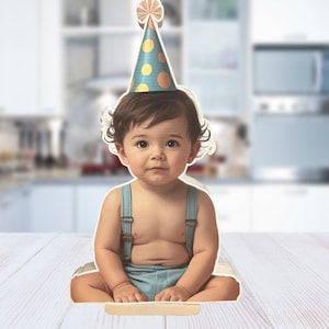 May include: A cut-out image of a baby wearing a party hat with orange and yellow accents and blue polka dots. The baby is wearing blue suspenders and shorts. The baby is sitting on a white surface.