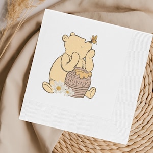 May include: White square paper napkin featuring a cartoon illustration of a yellow bear with a honey pot and a butterfly. The honey pot has the word "HUNNY" printed on it. Daisies are at the bottom of the bear.