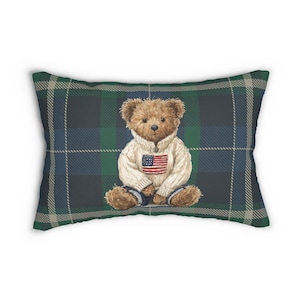 May include: Rectangular pillow with a plaid pattern in shades of green, blue, and white. The pillow features a teddy bear wearing a white sweater with an American flag on it. The teddy bear is sitting down.
