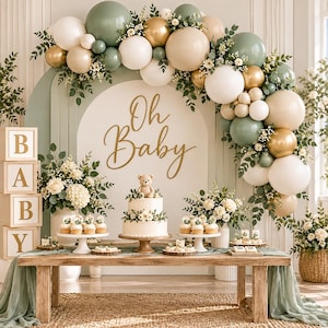 May include: A baby shower display featuring a wooden table with a two-tier cake topped with a teddy bear, cupcakes, and floral arrangements. An arch backdrop with sage green, white, and gold balloons displays the text Oh Baby, with stacked blocks.