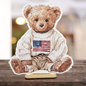 May include: A wooden cutout of a teddy bear wearing a white sweater with an American flag design. The bear is seated, with tan pants and brown fur. The cutout is mounted on a small wooden base, suitable for display.