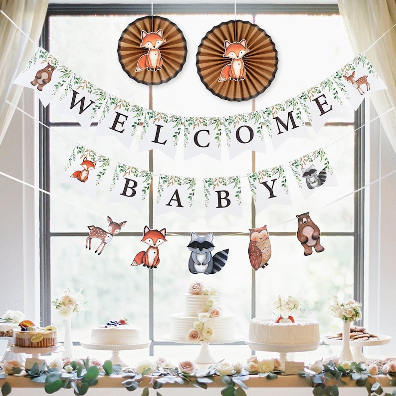 May include: Woodland themed baby shower decorations featuring a welcome banner, baby banner, and two paper fans with foxes. The banners are white with green leaves and black lettering. The fans are brown with foxes in the centre.