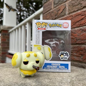 Könnte beinhalten: Eine gelbe und weiße Funko Pop! Vinylfigur eines Pokémon namens Fidough. Die Figur hält einen kleinen braunen Zigarren in ihrem Mund. Die Box ist mit "Pop! Pokemon 1051 Fidough Patachio Heifel" beschriftet.