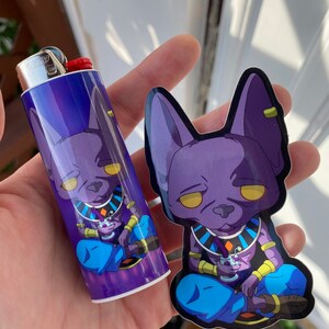 May include: A purple and blue lighter and sticker set featuring a cartoon character with yellow eyes. The lighter is a standard size, and the sticker is die-cut. The character is depicted in a seated position, holding a drink.