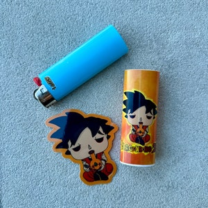 May include: A light blue BIC lighter, a sticker, and a cylindrical container. The sticker and container feature a cartoon character with black hair and red clothing, holding a smoking pipe. The container has the text "DRAGON BONG Z".