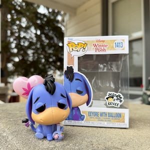 Puede incluir: Una figura de vinilo Funko Pop! de Ígor de Winnie the Pooh, sosteniendo un globo rosa en forma de corazón. La figura es azul con un hocico marrón y pelo negro. La caja dice "Eeyore with Balloon".