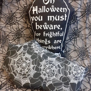 May include: Two black and white Halloween pillows. One pillow is shaped like a tombstone with the text "On Halloween you must beware, for frightful things are everywhere." The other pillow has a white spiderweb design on a black background.
