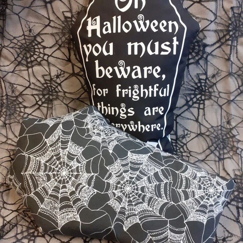 May include: Two black and white Halloween pillows. One pillow is shaped like a tombstone with the text "On Halloween you must beware, for frightful things are everywhere." The other pillow has a white spiderweb design on a black background.