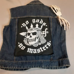 May include: A blue denim vest with a black patch on the back. The patch features a skull with a crown and the text "no gods - no masters".
