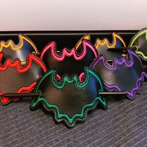 May include: Six black bat-shaped hair clips with colourful embroidered edges. The clips have a black stick attached to the back for holding hair.