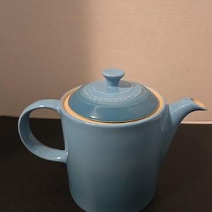 May include: A light blue ceramic teapot with a matching lid featuring the embossed text LE CREUSET. The teapot has a cylindrical body, a curved handle on the left side, and a short spout on the right side. The interior rim is a cream color.