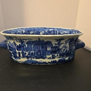 May include: An oval ceramic serving bowl featuring a blue and white transferware pattern depicting a historical landscape with figures, horses, and buildings. The bowl has a scalloped rim and two solid blue handles on opposite sides.