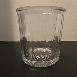 Op de afbeelding: Een helder glazen drinkglas met verticale gegroefde panelen langs de zijkanten en een zonnestraalpatroon in reliëf op de ronde basis. Het vat heeft een gladde, afgeronde rand en een cilindrische vorm met rechte wanden van dik, stevig glas.