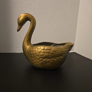 May include: A gold-colored swan-shaped planter or decorative bowl featuring a textured feather pattern on its body. The swan has a long, curved neck and a hollowed-out center, with a metallic finish that shows subtle variations in tone and texture.