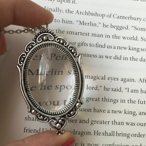 May include: A silver-toned magnifying glass necklace with a heart-shaped pendant. The magnifying glass has a clear lens and the text "Merlin's" is visible through the lens. The necklace is on a book with text about Merlin.