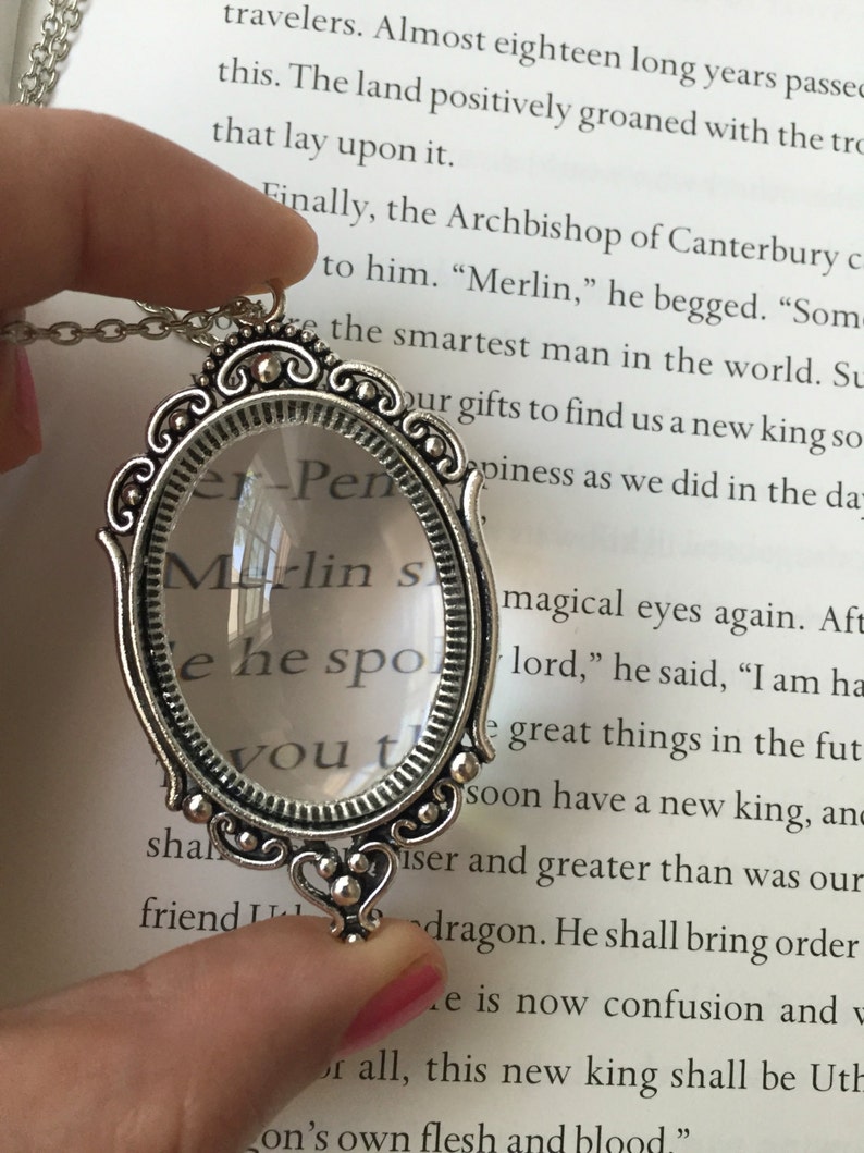 May include: A silver-toned magnifying glass necklace with a heart-shaped pendant. The magnifying glass has a clear lens and the text "Merlin's" is visible through the lens. The necklace is on a book with text about Merlin.