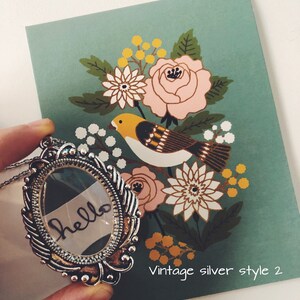 May include: A silver locket necklace with a glass insert that says "hello" in black ink. The locket is surrounded by an ornate silver frame. The necklace is on a green background with a floral pattern and a bird.
