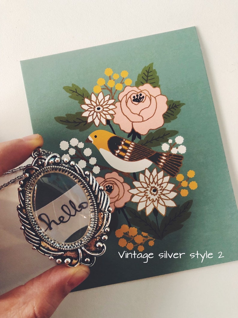 May include: A silver locket necklace with a glass insert that says "hello" in black ink. The locket is surrounded by an ornate silver frame. The necklace is on a green background with a floral pattern and a bird.
