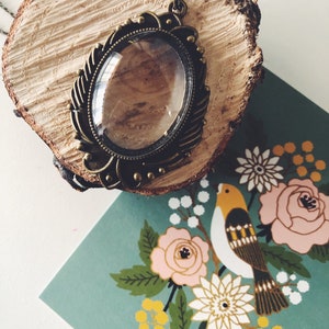 May include: A vintage gold-toned pendant necklace with a clear glass oval charm. The necklace is displayed on a wooden surface with a floral pattern background.
