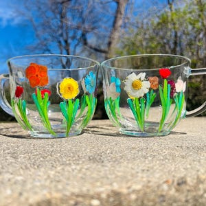 May include: Two clear glass teacups with handles, each adorned with hand-painted floral designs. The flowers include red, orange, yellow, white, and blue blooms with green stems and leaves. The cups are set on a concrete surface.