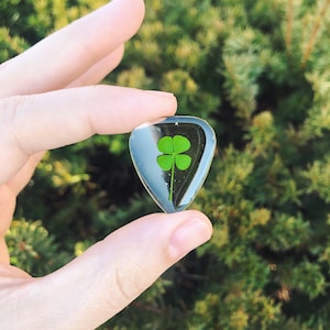 May include: A clear guitar pick with a four-leaf clover pressed inside. The clover is green and the pick is shaped like a teardrop.