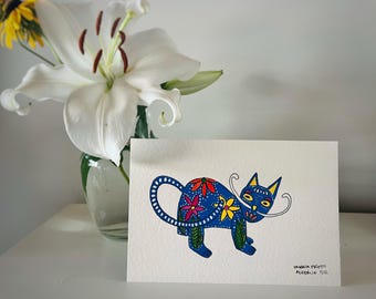 May include: A blue cat illustration with floral accents on a white card. The cat has a long tail and whiskers. A vase of white lilies and yellow flowers is in the background. The card is signed "Valeria Prieto, Alebrije 11/12."