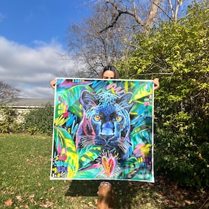 May include: A colorful painting of a black panther with a heart in the center, surrounded by green and yellow foliage. The painting is framed and held by a person.