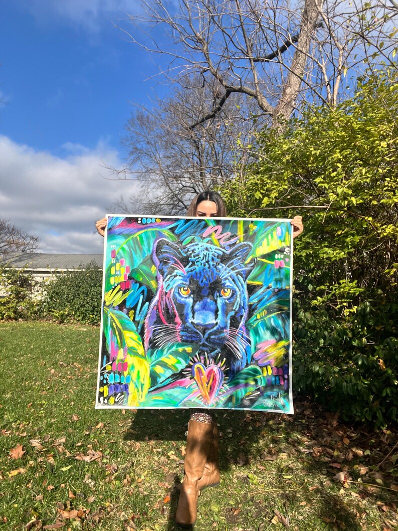May include: A colorful painting of a black panther with a heart in the center, surrounded by green and yellow foliage. The painting is framed and held by a person.