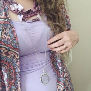 May include: A woman wearing a purple tank top with a layered necklace featuring a magnifying glass pendant. She is also wearing a colorful paisley patterned kimono and a ring on her finger.